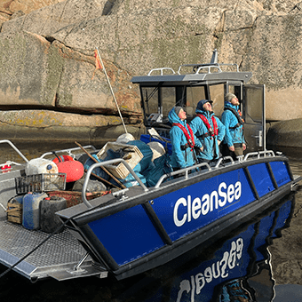 CleanSea-gänget, fyra av dem, står upp i den blå arbetsbåten. Vattnet ligger stilla.